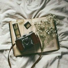 an open book with a camera and flowers on it sitting on top of a bed