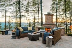 an outdoor fireplace surrounded by wicker furniture on a stone patio with pine trees in the background