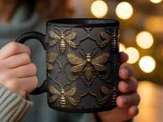 a woman holding a black and gold coffee mug with bees printed on it's side