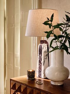 a white vase sitting on top of a wooden table next to a lamp with a green plant in it