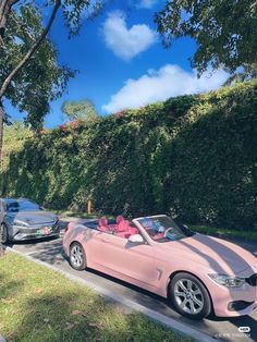 a pink convertible car driving down a street next to a hedge