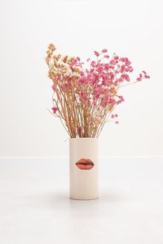 a white vase with pink flowers and lipstick on the lip is shown in front of a white wall