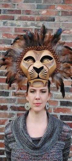 a woman wearing a lion mask with feathers on it's head, standing in front of a brick wall