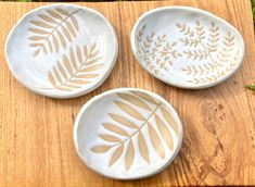 three white bowls with gold designs on them sitting on a wooden table next to grass