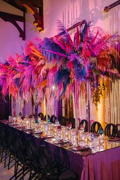 a long table topped with lots of tables covered in purple and pink feathers next to tall vases filled with candles