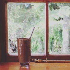 a glass filled with chocolate sitting on top of a wooden table next to a window