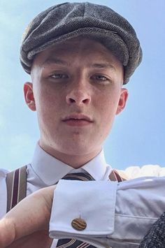 a young man wearing a hat and suspenders is holding his tie up to his chest