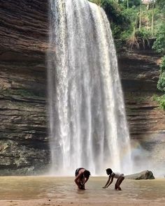 two people standing in front of a waterfall with a rainbow behind them and water falling off the side