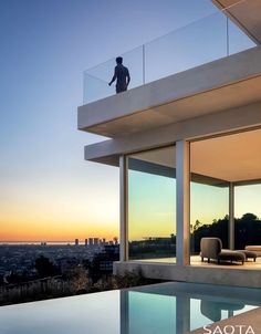 a man standing on the balcony of a modern house overlooking a pool and cityscape