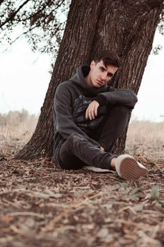 a young man sitting under a tree in the woods with his foot on the ground