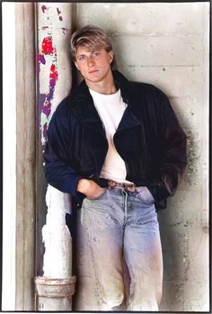 a young man leaning against a wall with his hands in his pockets and looking at the camera