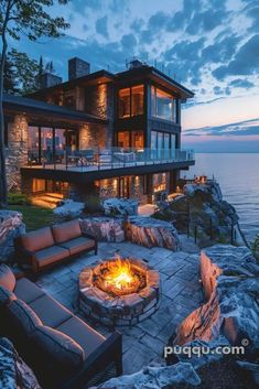 an outdoor fire pit in front of a large house by the water at night with lights on