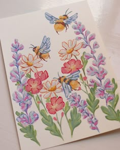 a card with watercolor flowers and bees on it, sitting on a white surface