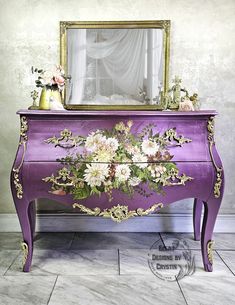 a purple dresser with flowers painted on it and a gold framed mirror in the background