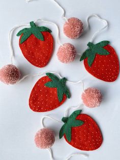 four strawberries with pom - poms hanging from string on white surface,