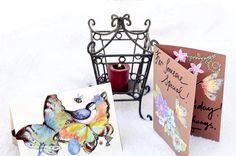 two cards with butterflies on them next to a candle and a lantern in the snow