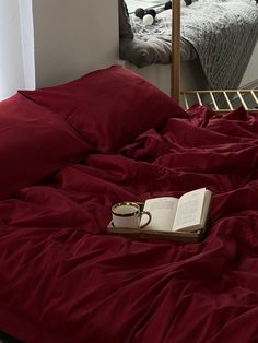 an open book on a bed with red sheets and pillows in front of a mirror