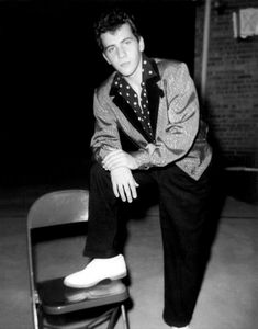 a man sitting on top of a chair next to a black and white photo with his legs crossed