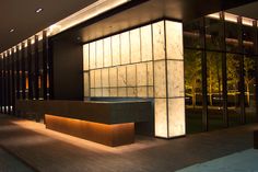 an empty bench in front of a building with glass walls and lights on the outside