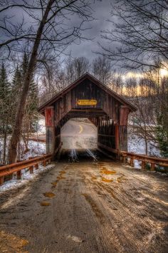 an old covered bridge in the winter with snow on the ground and trees around it