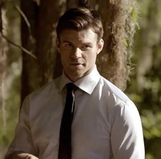 a man wearing a white shirt and black tie standing in front of a tree trunk