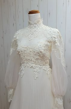 a white wedding dress on a mannequin with sheer sleeves and lace detailing, in front of a wooden background