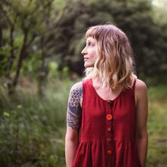 a woman in a red dress looking off into the distance with trees and bushes behind her