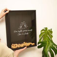 a woman holding up a framed sign with peanuts in front of her and a potted plant