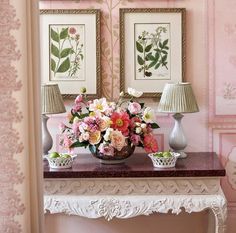 a vase with flowers on a table in front of pink wallpaper and framed pictures