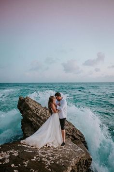 a bride and groom standing on rocks by the ocean with waves crashing in front of them