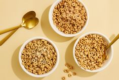 three bowls filled with cereal next to two spoons on a yellow table top,