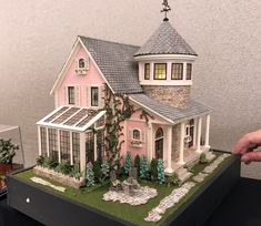 a pink doll house is displayed on a black table with green plants and flowers around it