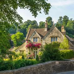 an old stone house with pink flowers on the front and side windows, surrounded by greenery