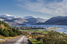 an empty road with mountains in the background and blue water on either side that is surrounded by trees