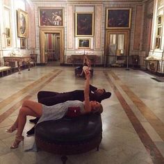 two women laying on top of a round ottoman in a room filled with paintings and chandeliers