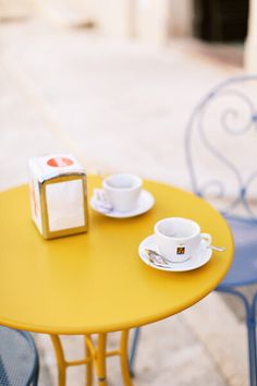 a yellow table with two white cups and saucers on it next to a blue chair