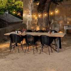 a wooden table with chairs around it under a tree and lights strung from the branches