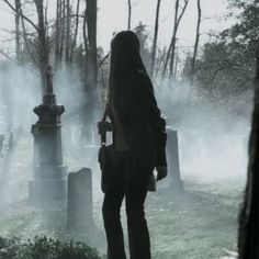 a person standing in front of a cemetery on a foggy day with trees and tombstones