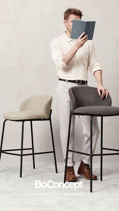 a man sitting on a chair reading a book with his hands in his mouth while standing next to two stools