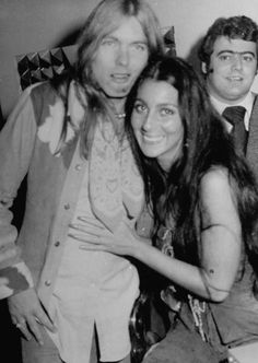 three people posing for the camera in front of a man with long hair and an unbuttoned shirt