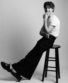 a man sitting on top of a wooden stool next to a chair with his legs crossed