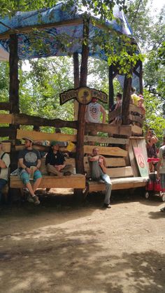 several people sitting on wooden benches in the shade under an umbrella covered area with trees