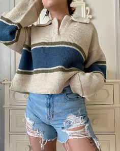 a woman wearing ripped shorts and a striped sweater poses for the camera with her hands on her head