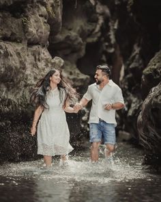 a man and woman are walking through the water in front of some rocks holding hands