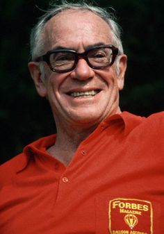 an older man wearing glasses and a red shirt smiles at the camera with his hand up in the air