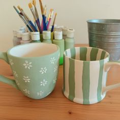 two coffee mugs sitting next to each other on top of a wooden table with paintbrushes