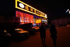 two people standing in front of a building with lights on it and cars parked outside