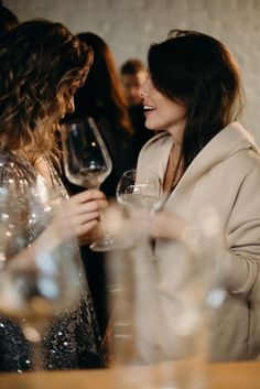 two women standing next to each other at a table with wine glasses in front of them