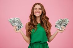 a woman holding two stacks of money in both hands and smiling at the camera while standing against a pink background