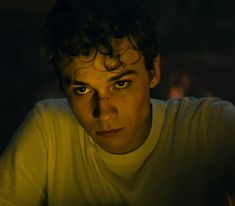 a young man with curly hair and green eyes stares at the camera in a dimly lit room